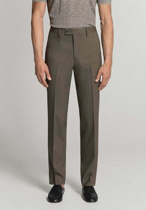 Hombre vestido con pantalones marrones ajustados, camiseta gris de manga corta y zapatos de vestir negros, de pie sobre un fondo neutro.