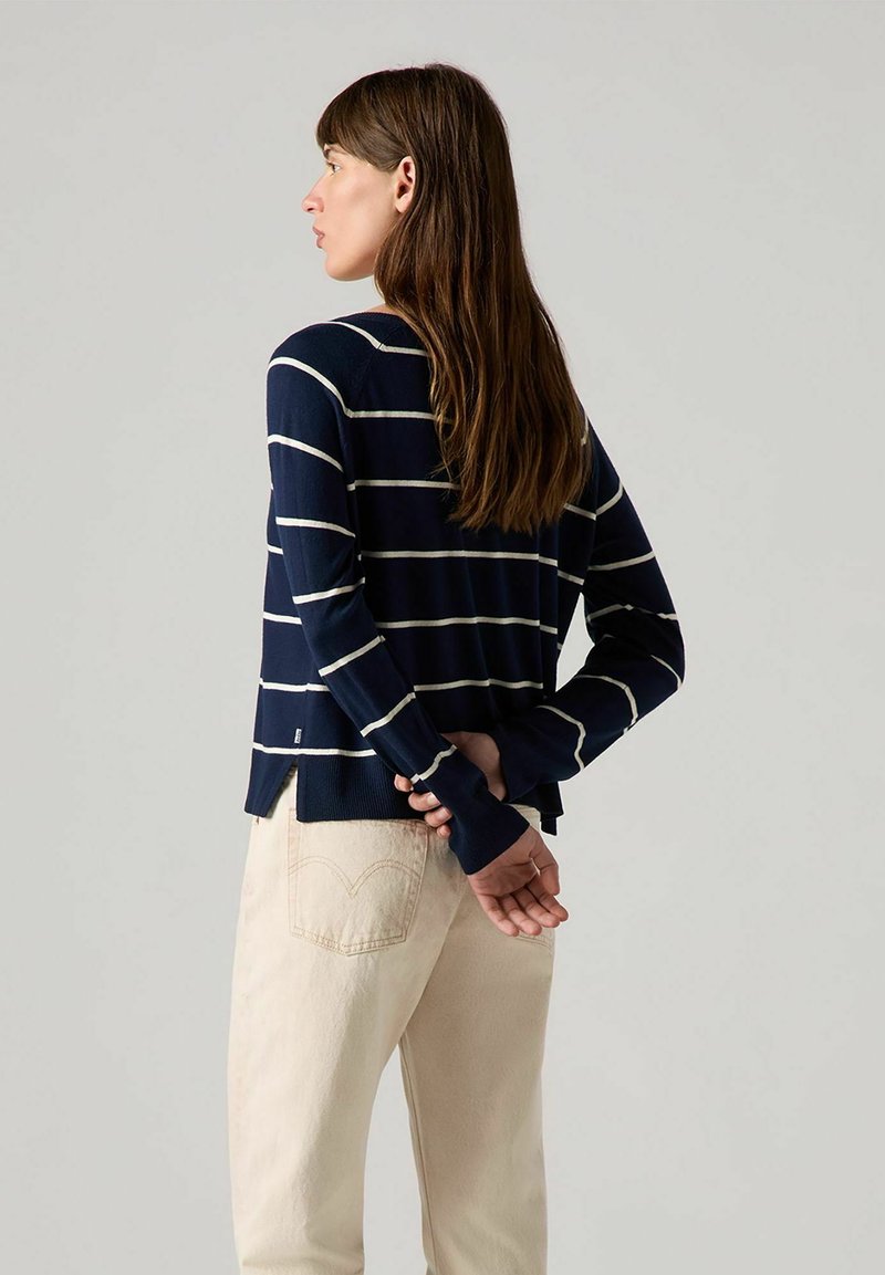Pull rayé marine avec des lignes crème, matière tricot légère, coupe décontractée, fentes latérales, porté avec un pantalon crème. Vue de dos.