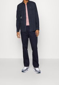 Tmavě modrá košile na knoflíky přes červeno-bíle pruhované tričko, v kombinaci s tmavě modrými kalhotami a vícebarevnými sportovními botami.