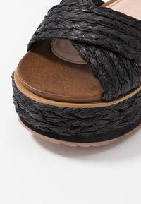 Svart flätad sandal med remmar och en brun fusk läder innersula samt en lagerplattform sula. Har en texturerad, vävd design.