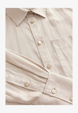 Gai bēšs pogu krekls, izgatavots no strukturētas auduma, ar apkakli, priekšējo kabatu un bēšām pogām ar maigu spīdumu.