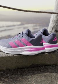 Baskets de sport grises avec des accents roses et un design à trois bandes. Tige en mesh texturé, semelle blanche rembourrée et logo visible sur le talon.