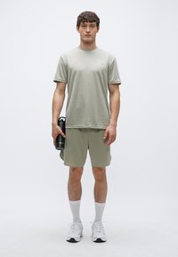 Tricou și pantaloni scurți atletici verde deschis, realizați din material care elimină transpirația. Modelul ține o sticlă de apă neagră, purtând șosete și pantofi atletici albi.