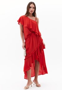 Abito rosso monospalla con strati arricciati e orlo asimmetrico. Realizzato in tessuto leggero. Abbinato a una pochette argentata e tacchi con cinturini.