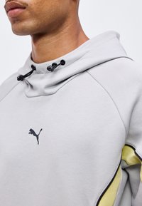 Gros plan d'une personne portant un sweat à capuche Puma gris clair avec des cordons noirs et des accents jaunes et noirs sur la manche.