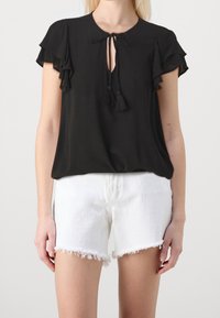 Frau trägt eine schwarze kurzärmlige Bluse mit Quastenbindemöglichkeiten und weiße ausgefranste Denim-Shorts vor einem einfarbigen Hintergrund.