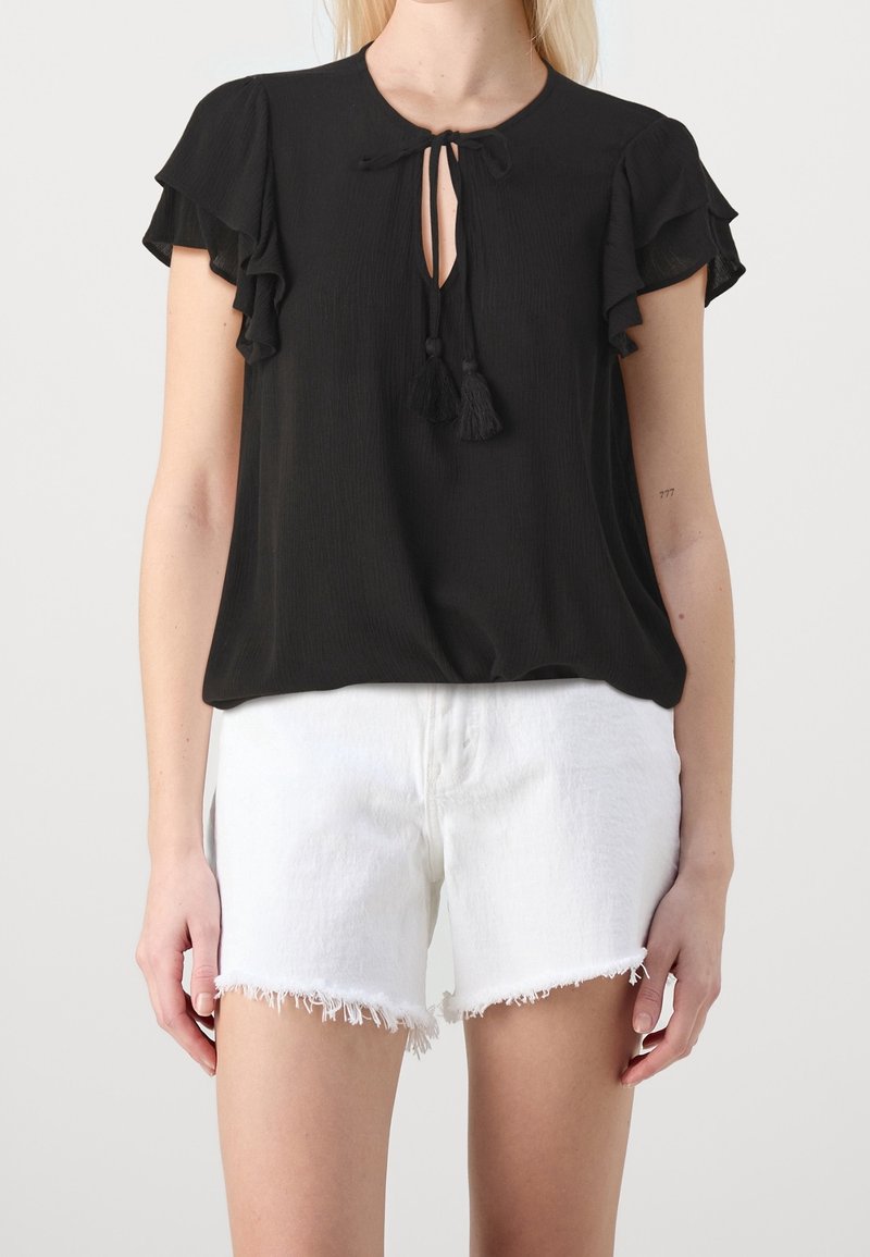 Donna che indossa una blusa nera a maniche corte con laccetti a frange e pantaloni shorts in denim bianchi sfilacciati su uno sfondo neutro.