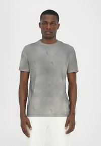 Grå kortærmet t-shirt med et diskret logo mønster over hele, rund hals og blød stofstruktur. Båret med hvide bukser.