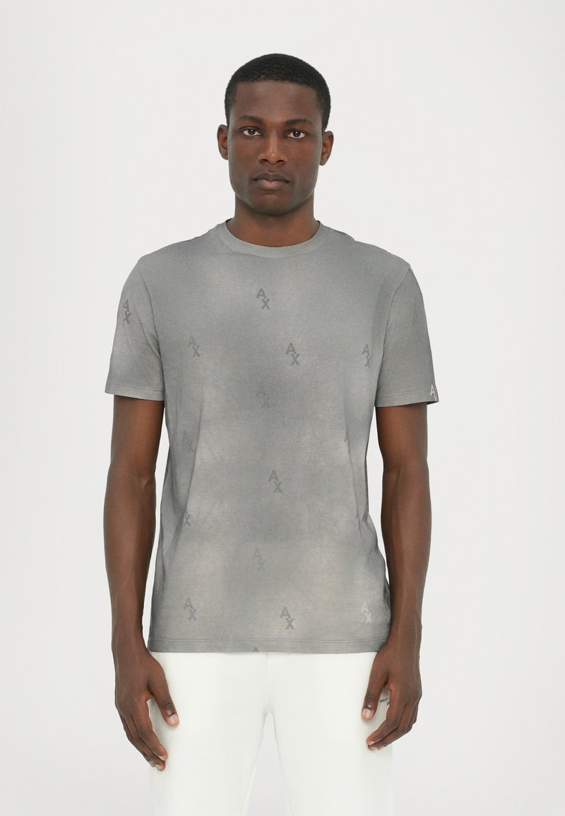 Grå kortærmet t-shirt med et diskret logo mønster over hele, rund hals og blød stofstruktur. Båret med hvide bukser.