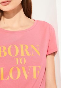 Lyserød bomulds t-shirt med korte ærmer, der har fed gul tekst, som siger "BORN TO LOVE." Glat tekstur og rund halsudskæring.