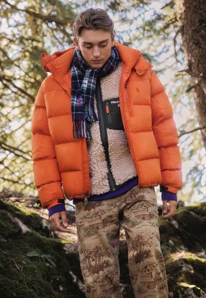 Un joven mira hacia abajo mientras está de pie en una zona boscosa. Lleva una chaqueta acolchada naranja, una bufanda de cuadros, un chaleco polar y unos pantalones estampados.