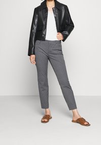 Schwarze, cropped Lederjacke, weißes V-Ausschnitt-Oberteil, grau-weiße gestreifte, figurbetonte Hose und braune Rutschsandalen. Glatte Texturen und maßgeschneiderte Passform.