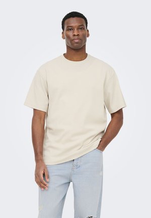 Homme portant un t-shirt crème côtelé à manches courtes et un jean bleu clair déchiré, debout devant un fond blanc uni.