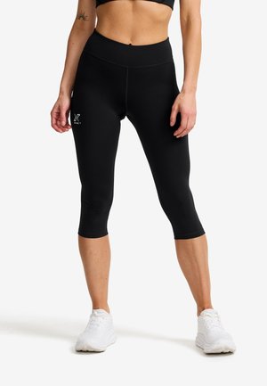 Kvinna klädd i svarta, korta träningsleggings och vita sneakers, stående med benen isär framför en vit bakgrund.