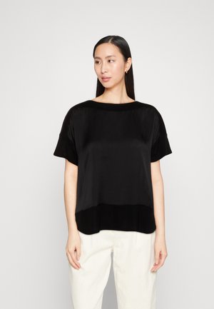 Μαύρο κοντομάνικο τοπ φτιαγμένο από απαλό ύφασμα, με χαλαρή εφαρμογή και στρογγυλή λαιμόκοψη, συνδυασμένο με μια ελαφρώς πιο μακριά λεπτομέρεια στο τελείωμα.