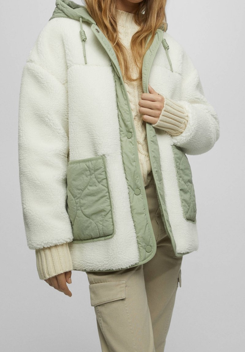 Personne portant une veste polaire blanche avec des poches matelassées vertes et des bordures, un pull en maille beige et un pantalon cargo beige.