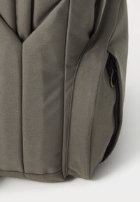 Sac à dos en tissu gris avec un design rembourré, présentant une surface texturée et une poche latérale avec une fermeture éclair noire.