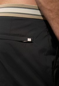 Un short noir présente une ceinture rayée beige et noire, avec une petite poche zippée à l'arrière, accentuée par un détail en métal argenté.