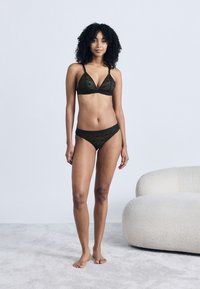 Γυναίκα με σγουρά μαλλιά, φορώντας μαύρο δαντελένιο σουτιέν και εσώρουχα, στέκεται ξυπόλυτη σε ανοιχτόχρωμο χαλί κοντά σε μια λευκή μοντέρνα καρέκλα με υφή.