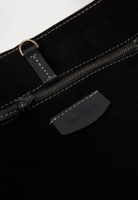 Mustan nubukilaukun, jossa on kontrastivärinen valkoinen ommel, vetoketjullinen suljenta, nahkavedin ja brändätty nahkamerkki.