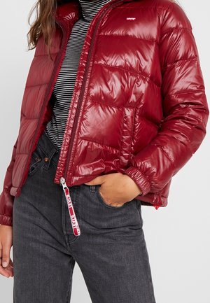 Personne portant une doudoune rouge brillante de Levi's par-dessus un col roulé à rayures noires et blanches, jean gris foncé avec la main dans la poche.