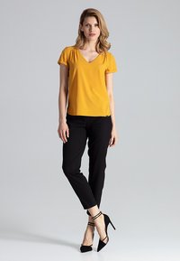 Gelbe kurzärmelige V-Ausschnitt-Bluse, kombiniert mit schwarzen maßgeschneiderten Hosen und schwarzen High Heels. Glattes Material mit einer figurbetonten Silhouette.