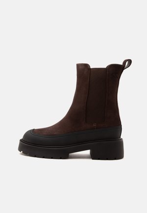 Bota de ante marrón hasta el tobillo con suela gruesa de goma negra, panel lateral elástico, lengüeta en el talón y diseño de puntera redonda.