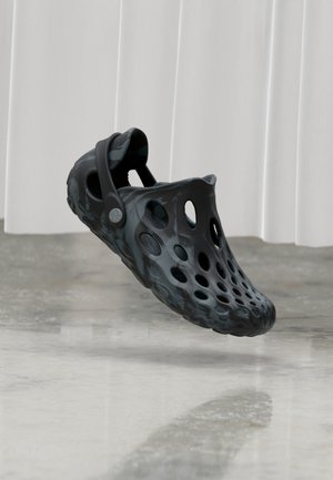 Crna slip-on cipela s višestrukim ventilacijskim otvorima i remenom na peti, visi iznad poliranog poda na bijeloj zavjesi u pozadini.