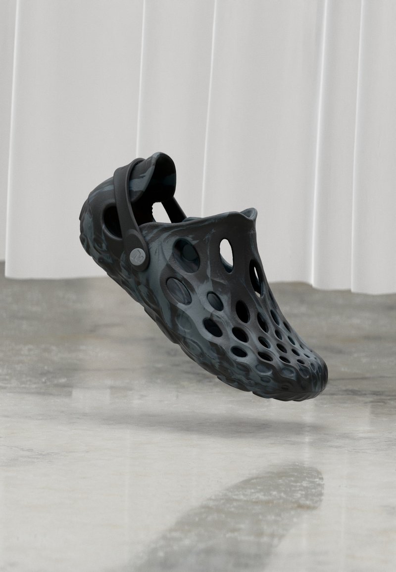 Chaussure noire à enfiler avec plusieurs trous de ventilation et une sangle au talon, suspendue au-dessus d'un sol poli devant un rideau blanc.