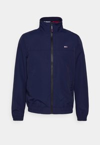 Námořnicky modrá bunda na zip z lehké tkaniny, s stojáčkovým límcem, elastickými manžetami a malým logem na levé straně hrudi.