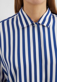 Chemise boutonnée à rayures bleu et blanche avec un col classique. Le tissu semble lisse ; les rayures sont bien espacées et vives.