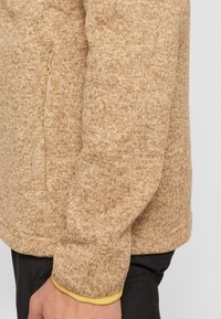 Pull en tricot beige avec un motif texturé, doté d'une poche latérale zippée et de détails jaunes contrastants aux poignets.