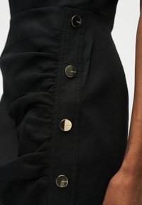 Κοντινό πλάνο μαύρου ρούχου από ραφτό ύφασμα με τέσσερα χρυσά κουμπιά με χαραγμένο το «PINKO» κατά μήκος της πλευράς κοντά στο μπράτσο ενός ατόμου.