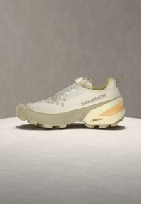 Μπεζ και εκρού παπούτσι για τρέξιμο σε μονοπάτια Salomon με ανθεκτική σόλα και αριθμητικό σχέδιο στο επάνω μέρος, εμφανίζεται σε φωτεινή κυκλική επιφάνεια.