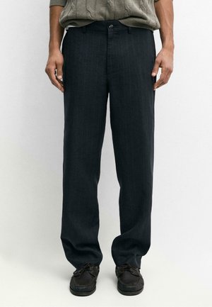 Mann trägt dunkel gestreifte Hose und dunkle Schnürschuhe, steht vor einem schlichten weißen Hintergrund.