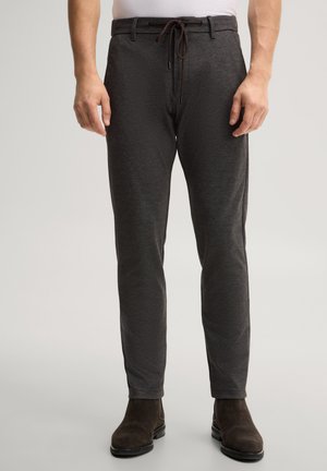 Mann trägt dunkelgraue, strukturierte Hose mit Tunnelzug in der Taille und dunkelbraune Wildlederstiefel vor neutralem Hintergrund.