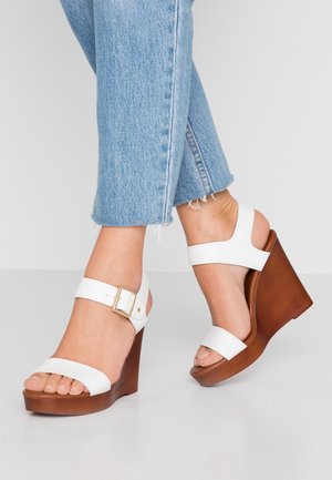 Fødder iført hvide kile-sandaler med brune træhæle og lyseblå kortere jeans på en ensfarvet lys baggrund.