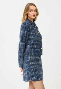 Młoda kobieta w niebiesko-białą kratkę kurtce z złotymi guzikami i pasującej spódnicy, stojąca bokiem na jednolitym tle.