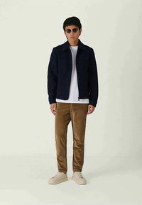 Homme portant des lunettes de soleil, une veste marine, un t-shirt blanc, un pantalon en velours côtelé marron et des baskets beige, se tenant devant un fond clair.