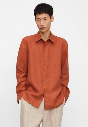 Mężczyzna w pomarańczowej, długorękawowej koszuli zapinanej na guziki i beżowych spodniach, stojący z rękami w kieszeniach na białym tle.