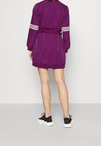 Lila idrottsklänning med lång ärm, vita randiga detaljer, elastisk midja och en rynkad design; i kombination med svartvita sneakers.