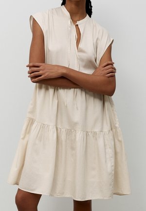 Kvinde iført en ærmeløs beige kjole med fine lodrette striber, stående med armene over kors mod en ensfarvet baggrund.