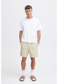 White cotton t-shirt, beige shorts with drawstring, white socks, and tan sandals. Model stands with hands in pockets against a plain background.