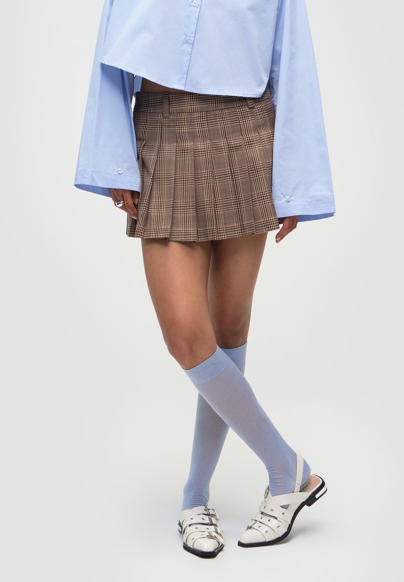 Gonna mini a pieghe a quadretti marroni, camicia azzurra a maniche lunghe, calze fino al ginocchio azzurre e sandali bianchi con accenti argentati.