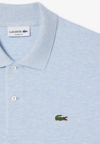 Ljusblå polo t-shirt i texturerad tyg, med klassisk krage, tre knappar fram och en broderad grön krokodil-logga.