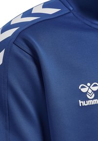 Maglietta atletica blu con texture liscia, caratterizzata da un motivo a chevron bianco sulla spalla e un logo sul petto. Il tessuto sembra leggero e elastico.