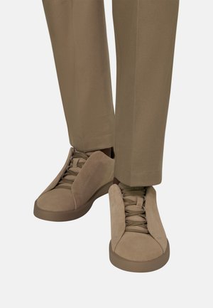 Beige Wildledersneaker mit breiten vorderen Klappen, die nach außen geöffnet sind, kombiniert mit passenden beigen Hosen, dargestellt vor weißem Hintergrund.