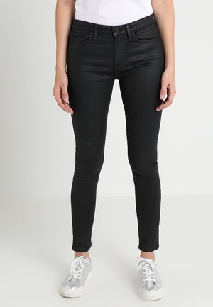 Femme portant un jean skinny noir et des baskets argentées, debout devant un fond clair uni.