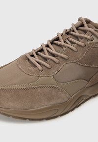Brown athletic shoes featuring a mix of textured suede and smooth fabric, with prominent stitching and flexible rubber soles. Laces in matching hue.
