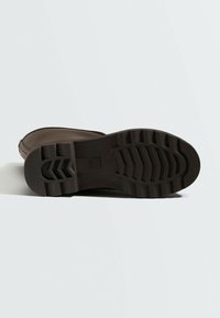 Semelle d’une botte en caoutchouc brun foncé avec des motifs de semelle profonds pour l’adhérence, montrée couchée sur le côté sur un fond clair uni.